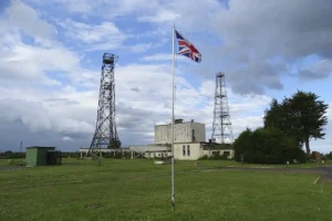 A view of the communication towers along with some of the buildings on site with the flag in the foreground.